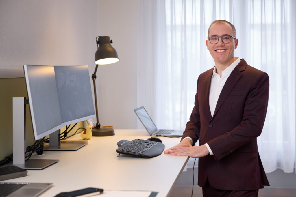Smiling man in a burgundy suit standing at a desk with dual computer monitors in a bright office.