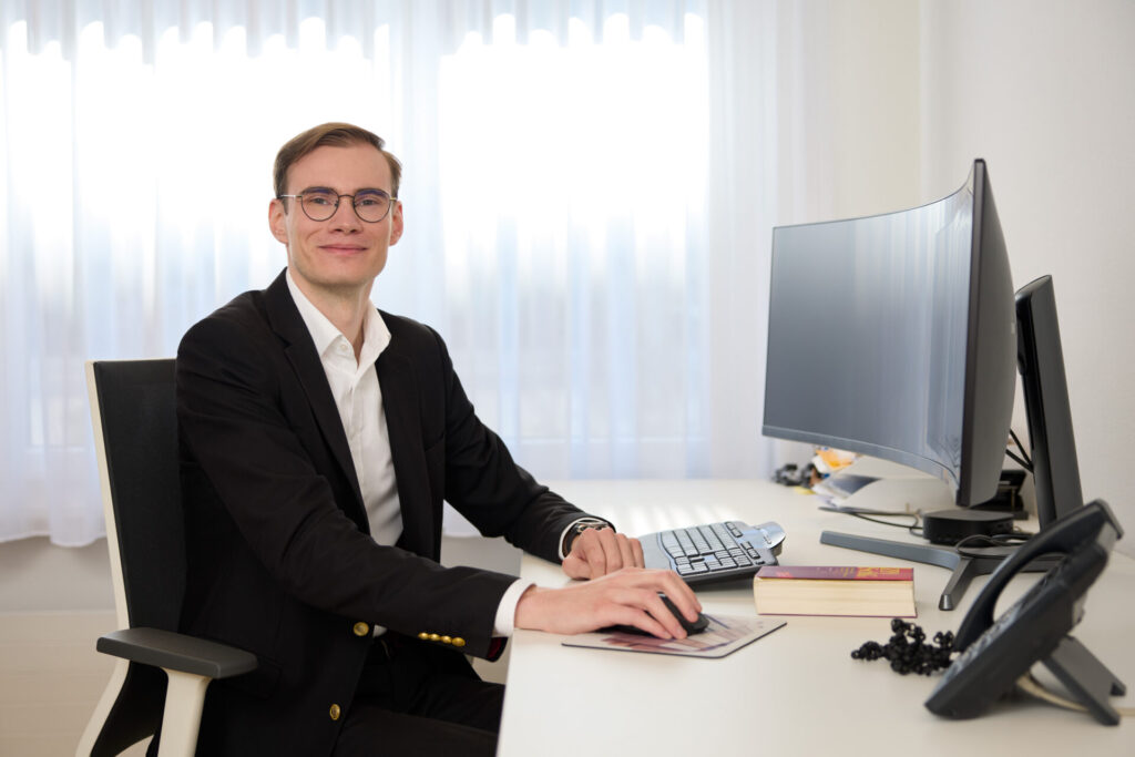 Professional man in a black suit sits at a white desk, typing on a keyboard with a curved monitor in front of him.