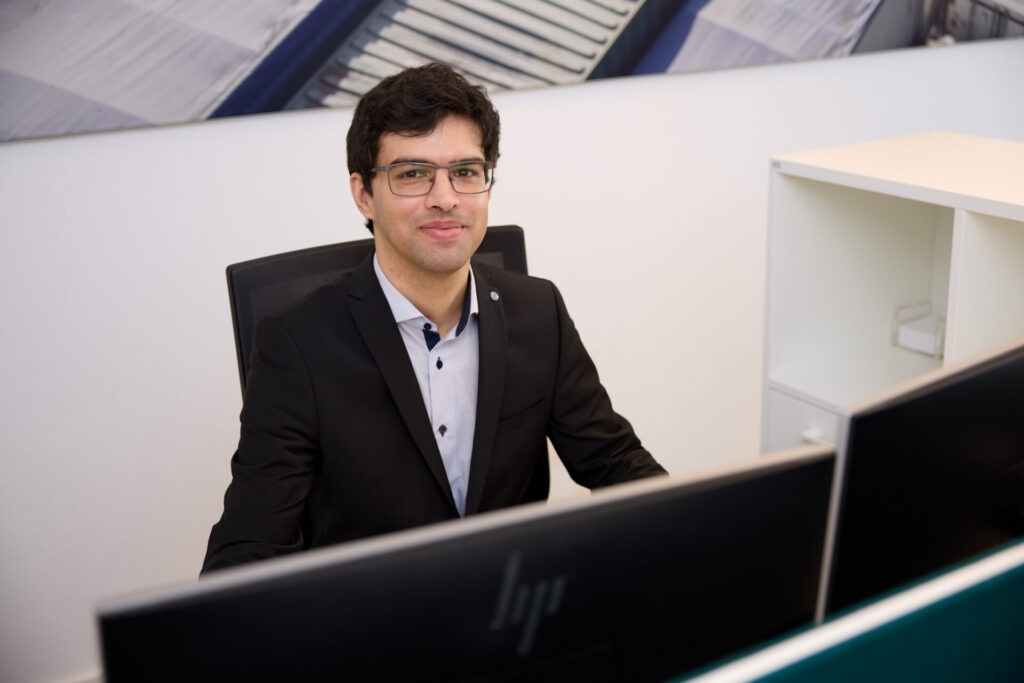 Man in a black suit and glasses sits at a desk with two computer monitors in a modern office.