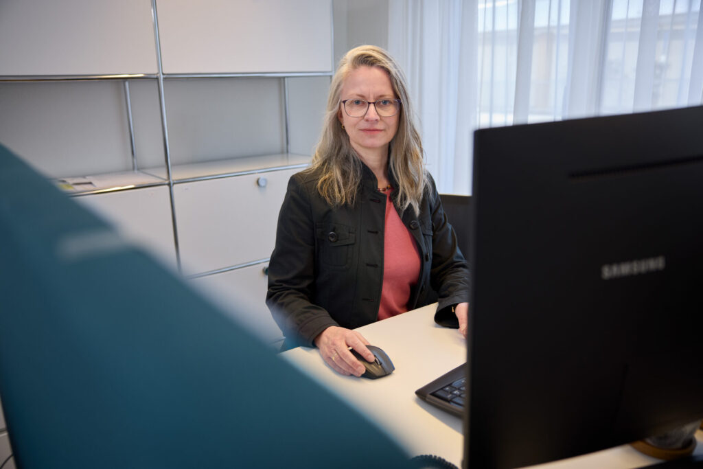 Woman with glasses sits at a desk using a computer mouse in an office setting, facing the camera.