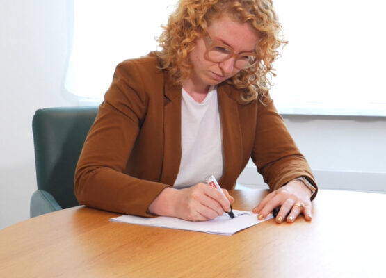 Woman with curly blond hair and glasses sits at a round wooden table, writing on paper with a red marker in a brown blazer.